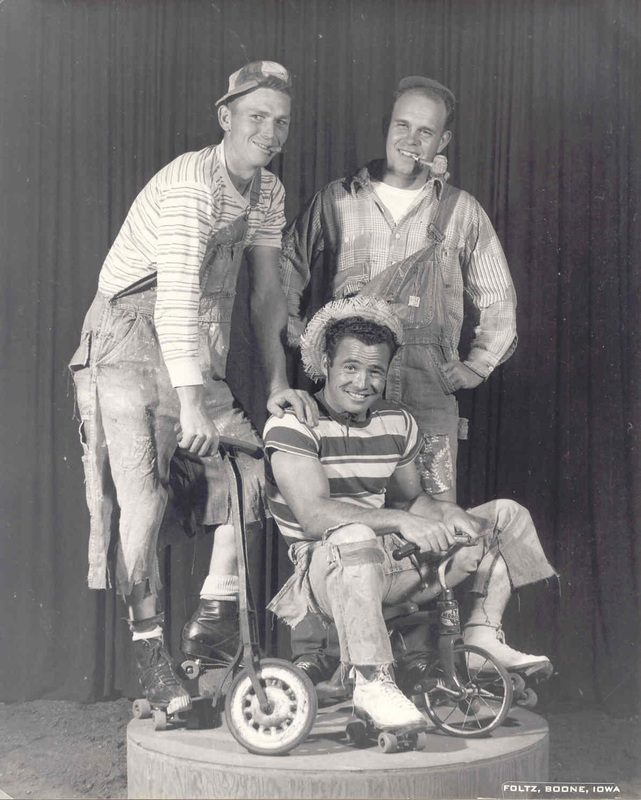 Three men posed with a seated man in a wheelchair; two men stand behind him in front of a dark curtain.